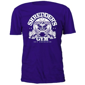Könnte beinhalten: Lilafarbenes T-Shirt mit einer weißen Grafik von Shredder aus Teenage Mutant Ninja Turtles, der Gewichte hebt. Die Grafik lautet "Shredder's Gym" und "Get Shredded".