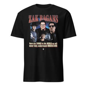 以下が含まれることがあります： 黒いTシャツ。4人の男性のグラフィックと、赤と白の「ZAK BAGANS」の文字がプリントされています。画像の下には「there are THINGS in this WORLD we will never fully understand UNDERSTAND」というテキストがあります。