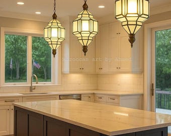 May include: Three pendant lights with geometric, cream-colored shades hang above a kitchen island. The shades are framed in a dark metal, and the island has a dark brown base with a light-colored countertop. The text "Moroccan art by Ahmed" is visible.
