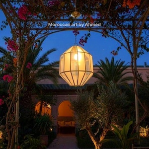 Op de afbeelding: Een buitenscène in de schemering toont een hangende geometrische lantaarn met een warme gloed. De lantaarn hangt aan een decoratieve metalen structuur met bloeiende wijnranken. De tekst "Moroccan art by Ahmed" is zichtbaar.