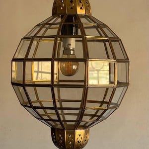 May include: A hanging, geometric pendant light with a brass frame and clear glass panels. The light fixture has a bulb inside and is suspended from a brass fixture. The text "Moroccan Art by Ahmed" is visible.