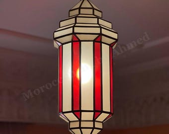 May include: Hanging Moroccan-style lantern with a geometric design. The lantern features alternating vertical panels of red and white, framed by black lines. The light fixture is suspended from a chain and has a pointed bottom.
