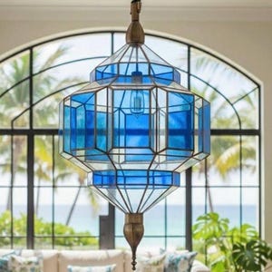 Può includere: Una lampada a sospensione in stile marocchino con un design geometrico. La lampada presenta pannelli in vetro colorato blu incorniciati da un metallo color bronzo. Il testo "Moroccan art by Ahmed" è visibile in alto. La lampada è appesa davanti a una grande finestra.