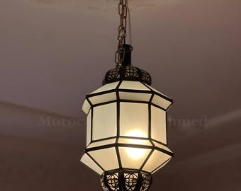 May include: A hanging lantern with a geometric design. The lantern has a brass-colored chain and frame, with white glass panels. The bottom of the lantern has an ornate, decorative pattern. The text "Moroc" is visible on the side.