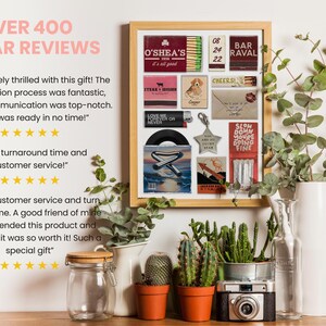 May include: A framed collage of matchboxes, a record, and other small decorative items. The matchboxes feature various designs and text, including "O'Shea's", "Bar Ravel", and "Cheers". The record is a 45 RPM record with a silver star on the cover. The collage is surrounded by a wooden frame and is displayed on a white wall with plants in front of it.