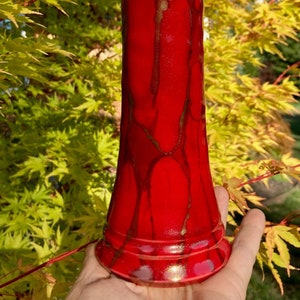 Puede incluir: Un jarrón de cerámica rojo con detalles dorados. El jarrón tiene una base acampanada y un cuerpo cilíndrico. Los detalles dorados están en forma de rayas y remolinos.