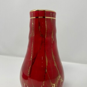 Puede incluir: Un jarrón de cerámica rojo con un borde dorado y líneas pintadas en oro. El jarrón tiene un cuerpo en forma de pera.