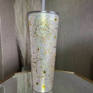 May include: A white tumbler with a gold glitter finish and a gold sun, moon, and stars design. The tumbler has a clear lid and a clear straw.