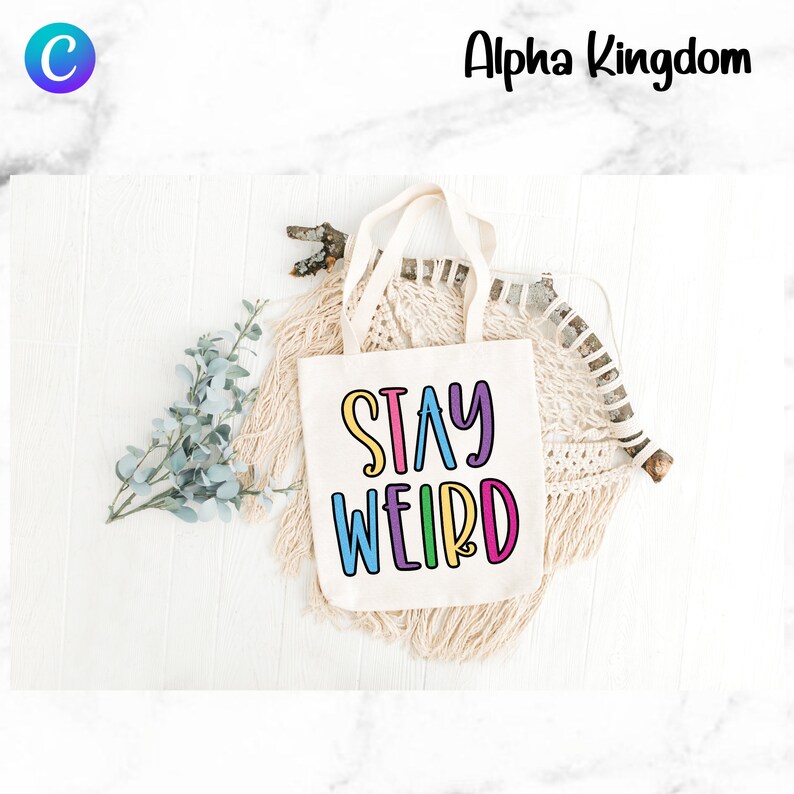 May include: A white canvas tote bag with the phrase "Stay Weird" printed in colorful letters. The bag is sitting on a white wooden surface with a branch and a macrame wall hanging in the background.