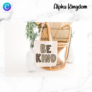 May include: A white tote bag with the words "BE KIND" in brown and beige letters. The bag is sitting on a wicker chair with green plants in the background.