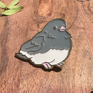 May include: A gray and white enamel pin of a cartoon bird with a pink beak and feet. The bird is sitting on a wooden surface.