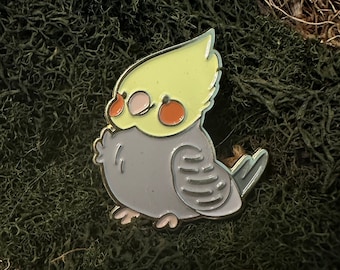 May include: A yellow and gray enamel pin of a cockatiel bird with red cheeks. The bird is facing left and has its wings tucked in. The pin is on a green mossy surface.