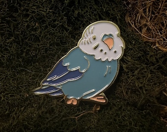 Può includere: Uno spillo in smalto blu e bianco con un piccolo uccello con un corpo blu chiaro, una testa bianca e un becco rosa. L'uccello è appollaiato su una superficie di muschio verde.