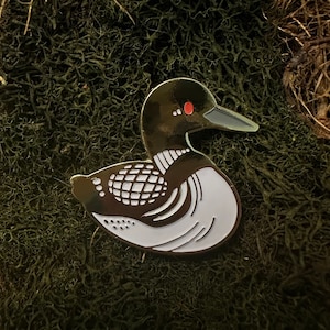 Puede incluir: Un pin de esmalte dorado de un colimbo, un tipo de ave buceadora, con marcas blancas y negras. El colimbo tiene un ojo rojo y un pico largo y puntiagudo.