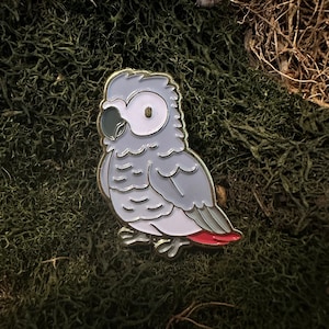 Puede incluir: Un pin de esmalte gris y blanco de un loro gris con cola roja. El loro está posado en una rama con las alas extendidas.