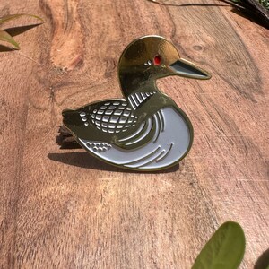 May include: A gold-coloured metal pin of a loon, a type of bird, with white detailing and red eyes. The pin is on a wooden surface.