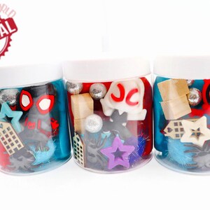 May include: Three clear jars filled with colorful play dough and various small toys, including a red and black Spiderman figure, a purple star, and a wooden building block. The jars have white lids and are labeled with the letters "JC".
