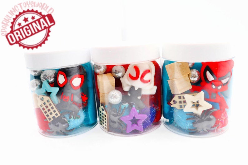 May include: Three clear jars filled with colorful play dough and various small toys, including a red and black Spiderman figure, a purple star, and a wooden building block. The jars have white lids and are labeled with the letters "JC".