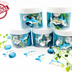May include: Five clear plastic jars with white lids filled with blue liquid, silver balls, and toy sharks. Additional toy sharks and blue glass beads are scattered around the jars. The jars are decorated with green leafy sprigs. The text "ORIGINAL" is in a red circle.