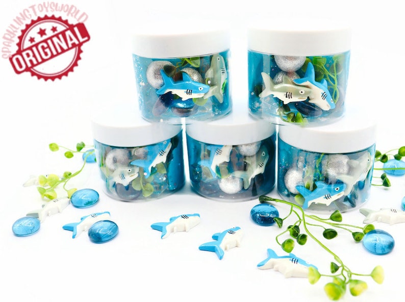 May include: Five clear plastic jars with white lids filled with blue liquid, silver balls, and toy sharks. Additional toy sharks and blue glass beads are scattered around the jars. The jars are decorated with green leafy sprigs. The text "ORIGINAL" is in a red circle.