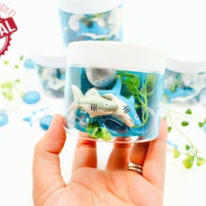 May include: Clear plastic jars filled with blue liquid, toy sharks, and greenery. The jars have white lids and are decorated with blue and white accents. The image includes the text "ORIGINAL" in a red circle.