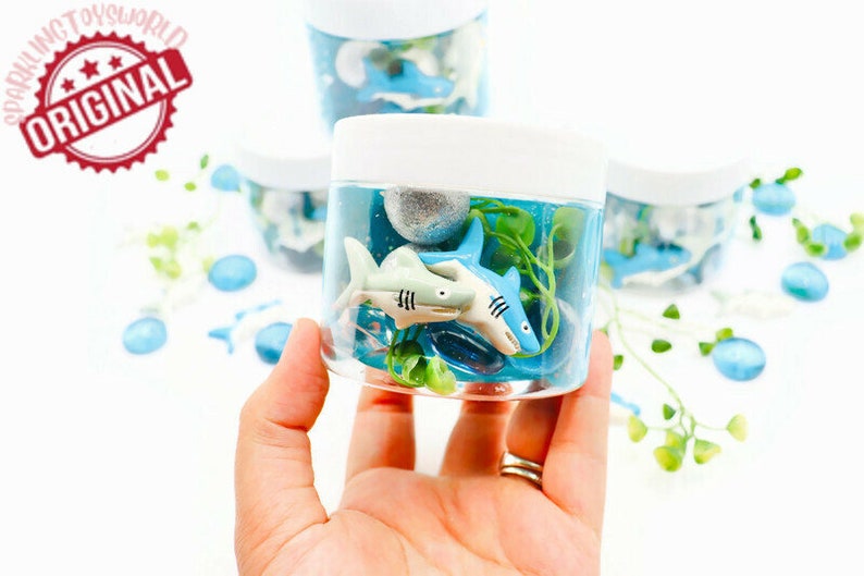 May include: Clear plastic jars filled with blue liquid, toy sharks, and greenery. The jars have white lids and are decorated with blue and white accents. The image includes the text "ORIGINAL" in a red circle.