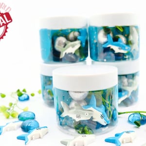 May include: Clear plastic jars with white lids filled with blue liquid, toy sharks, and decorative stones. The jars are stacked, with additional sharks and stones scattered around. The image includes the text "SPARKLING TOYSWORLD ORIGINAL".