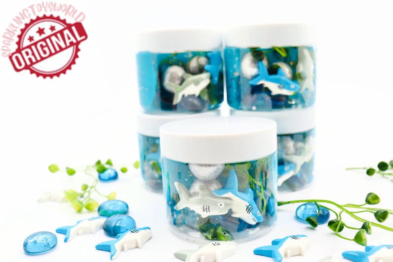 May include: Clear plastic jars with white lids filled with blue liquid, toy sharks, and decorative stones. The jars are stacked, with additional sharks and stones scattered around. The image includes the text "SPARKLING TOYSWORLD ORIGINAL".