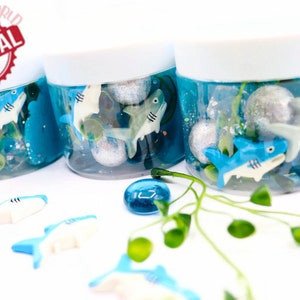 May include: Three clear jars filled with blue liquid, toy sharks, and decorative elements. The jars have white lids and are arranged on a white surface with scattered shark toys and blue glass beads. The image includes a red and white "ORIGINAL" stamp.