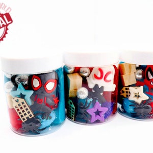 May include: Three clear plastic jars filled with colorful toy charms and beads. The jars have white lids. The charms include a red and black Spiderman figure, a white ghost with the letters "JC" on it, and a red and black Spiderman figure.