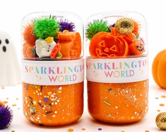 Peut inclure: Deux récipients en plastique transparent remplis de slime orange, de paillettes et de breloques sur le thème d'Halloween. Chaque récipient porte une étiquette blanche avec le texte SPARKLINGTO WORLD et des pompons verts, violets et orange.