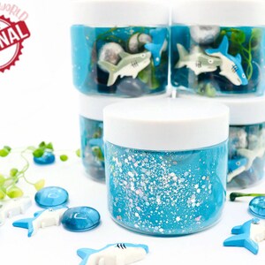May include: A collection of blue slime jars with white lids, decorated with shark figurines, stones, and aquatic plants. The jars are filled with blue glitter slime. The image includes the text "SPARKLING TOYSWORLD ORIGINAL".