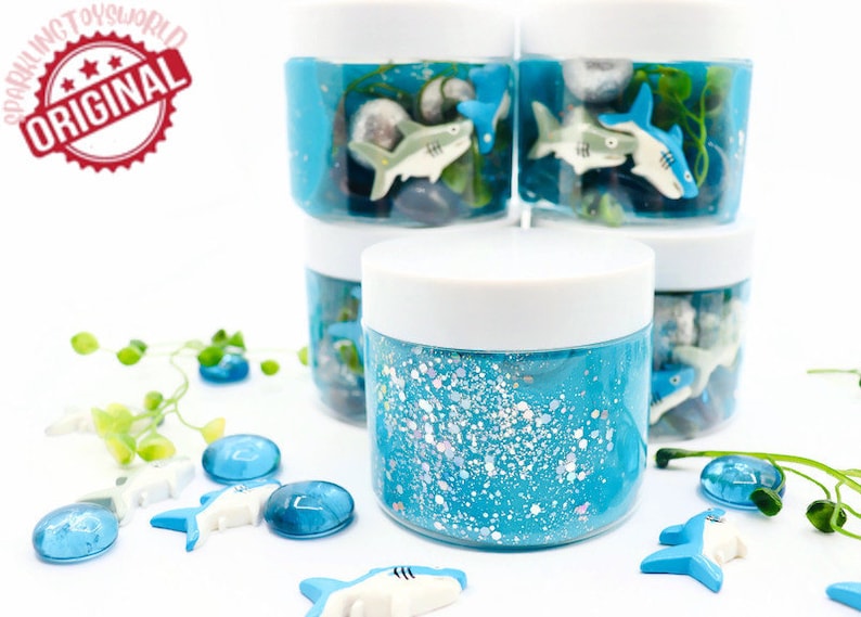 May include: A collection of blue slime jars with white lids, decorated with shark figurines, stones, and aquatic plants. The jars are filled with blue glitter slime. The image includes the text "SPARKLING TOYSWORLD ORIGINAL".