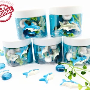 May include: Five clear plastic jars filled with blue liquid, silver spheres, and toy sharks. The jars have white lids and are arranged on a white surface. Scattered around are additional toy sharks and blue glass beads. The image includes a red and white "ORIGINAL" stamp.