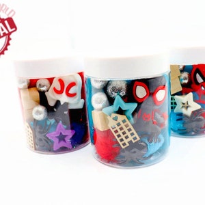May include: Three clear plastic jars with white lids filled with colorful plastic toys and trinkets. The jars contain a variety of shapes, including stars, hearts, and letters. The jars are arranged in a row on a white surface.