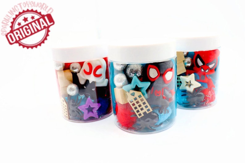 May include: Three clear plastic jars with white lids filled with colorful plastic toys and trinkets. The jars contain a variety of shapes, including stars, hearts, and letters. The jars are arranged in a row on a white surface.