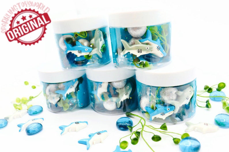 May include: Several clear plastic jars with white lids, filled with blue liquid, toy sharks, and decorative elements. The jars are arranged in a stack. The image includes the text "SPARKLING TOYSWORLD ORIGINAL".