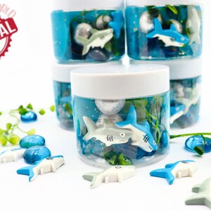 May include: Several clear plastic jars filled with blue liquid, small rocks, and toy sharks. The jars have white lids and are arranged in a stack. Additional toy sharks and blue glass beads are scattered around the jars. The image includes a red and white "ORIGINAL" stamp.