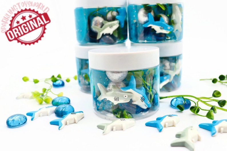 May include: Several clear plastic jars filled with blue liquid, small rocks, and toy sharks. The jars have white lids and are arranged in a stack. Additional toy sharks and blue glass beads are scattered around the jars. The image includes a red and white "ORIGINAL" stamp.