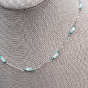 May include: A delicate necklace featuring a silver chain with alternating white and turquoise-colored beads. The necklace is displayed on a neutral-toned mannequin, showcasing its design and color contrast. The beads are cylindrical and evenly spaced.