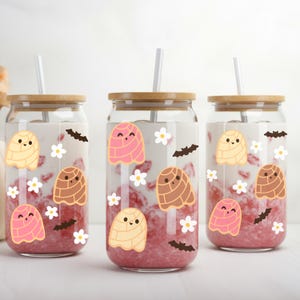 May include: Three glass tumblers with wooden lids and straws, filled with a pink and white beverage. Each tumbler is decorated with cartoon concha bread, white flowers, and bat stickers. The tumblers are perfect for cold drinks.