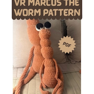 May include: Two orange crocheted worm figures with long limbs and large black and white eyes. The text VR MARCUS THE WORM PATTERN is displayed at the top in a brown scalloped banner, and a star-shaped badge with the text PDF is on the right.