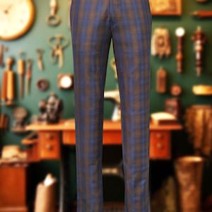 May include: Blue and brown plaid trousers displayed on a mannequin. The pants have a classic, tailored fit with a button closure and belt loops. The plaid pattern features vertical stripes of blue and brown. The background is a blurred workshop setting.