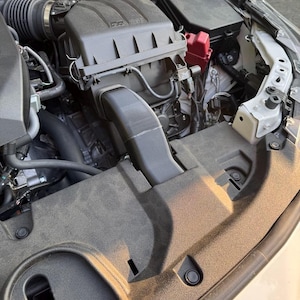 K&ouml;nnte beinhalten: Offener Motorraum eines Autos mit verschiedenen Komponenten. Der Motorraum zeigt einen schwarzen Luftfilterkasten, einen roten Sicherungskasten sowie mehrere Schl&auml;uche und Kabel. Der Motorraum ist staubig, mit einer schwarzen Kunststoffabdeckung im Vordergrund.