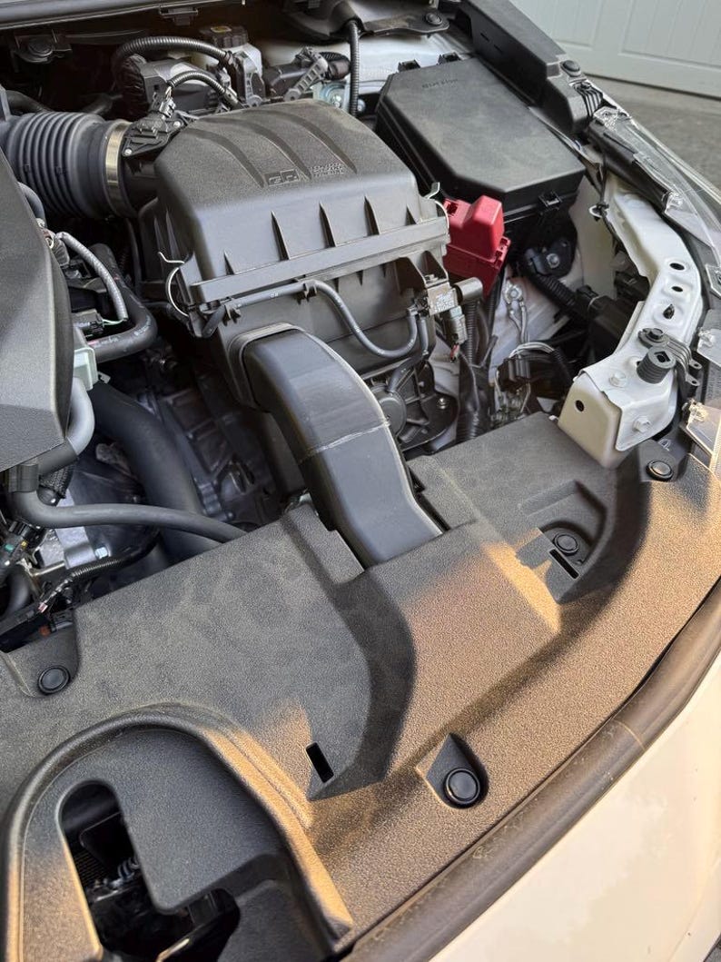 K&ouml;nnte beinhalten: Offener Motorraum eines Autos mit verschiedenen Komponenten. Der Motorraum zeigt einen schwarzen Luftfilterkasten, einen roten Sicherungskasten sowie mehrere Schl&auml;uche und Kabel. Der Motorraum ist staubig, mit einer schwarzen Kunststoffabdeckung im Vordergrund.