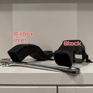 K&ouml;nnte beinhalten: Schwarzer Lufteinlass und serienm&auml;&szlig;ige Luftansaugkomponenten mit einem Schraubenschl&uuml;ssel. Der Lufteinlass ist gebogen und rechteckig. Der Text "Airbox Inlet" ist rot. Das Wort "Stock" ist rot auf dem serienm&auml;&szlig;igen Lufteinlass.