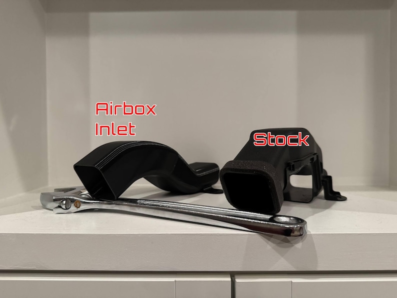 K&ouml;nnte beinhalten: Schwarzer Lufteinlass und serienm&auml;&szlig;ige Luftansaugkomponenten mit einem Schraubenschl&uuml;ssel. Der Lufteinlass ist gebogen und rechteckig. Der Text "Airbox Inlet" ist rot. Das Wort "Stock" ist rot auf dem serienm&auml;&szlig;igen Lufteinlass.