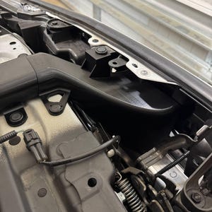 K&ouml;nnte beinhalten: Detaillierte Ansicht des Motorraums eines Autos, die verschiedene Komponenten zeigt. Das Bild zeigt schwarze Kunststoffteile, Metallhalterungen und Kabel, wobei der Schwerpunkt auf den komplizierten mechanischen Details liegt. Das Farbschema wird von Schwarz, Grau und Silber dominiert.
