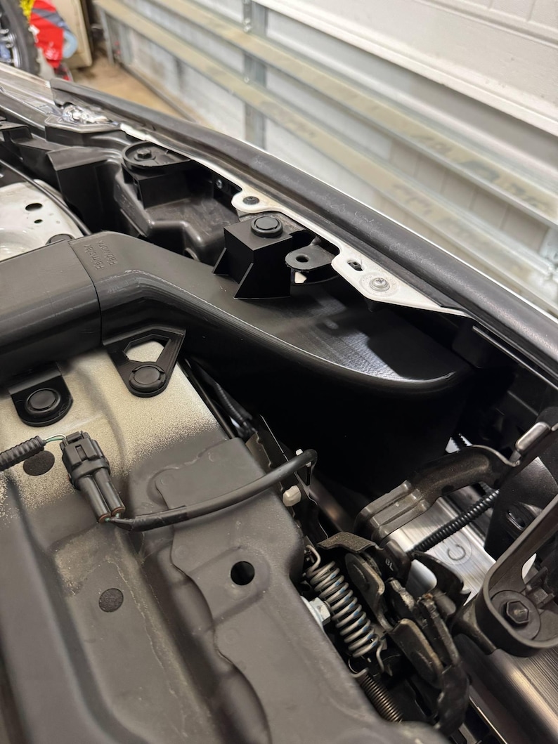 K&ouml;nnte beinhalten: Detaillierte Ansicht des Motorraums eines Autos, die verschiedene Komponenten zeigt. Das Bild zeigt schwarze Kunststoffteile, Metallhalterungen und Kabel, wobei der Schwerpunkt auf den komplizierten mechanischen Details liegt. Das Farbschema wird von Schwarz, Grau und Silber dominiert.