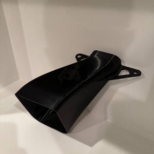 K&ouml;nnte beinhalten: Ein schwarzes, 3D-gedrucktes Objekt mit einer ausgestellten, rechteckigen &Ouml;ffnung und einem sich verj&uuml;ngenden K&ouml;rper. Es verf&uuml;gt &uuml;ber zwei Befestigungslaschen mit L&ouml;chern und ein kleines rechteckiges Detail oben. Das Objekt besteht aus mattem, schwarzem Material.